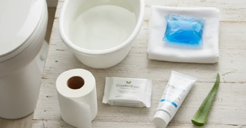 An organized flat lay of at-home remedies for hemorrhoid relief on a light wooden surface, including a steaming sitz bath, a cold pack on a towel, soft toilet paper, medicated wipes, and a tube of soothing cream.